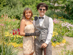 Two people in 1920s attire standing in a garden with flowers and trees in the background.