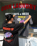 Person holding a t-shirt with 'WASHED UP' branding in front of a coin laundry sign.