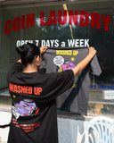 Person holding a t-shirt with 'WASHED UP' branding in front of a coin laundry sign.