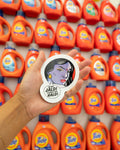 Hand holding a circular sticker with a character design in front of Tide laundry detergent bottles.