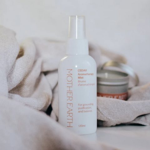 Mother Earth Cedar Aromatherapy Mist bottle with a candle on a soft fabric background